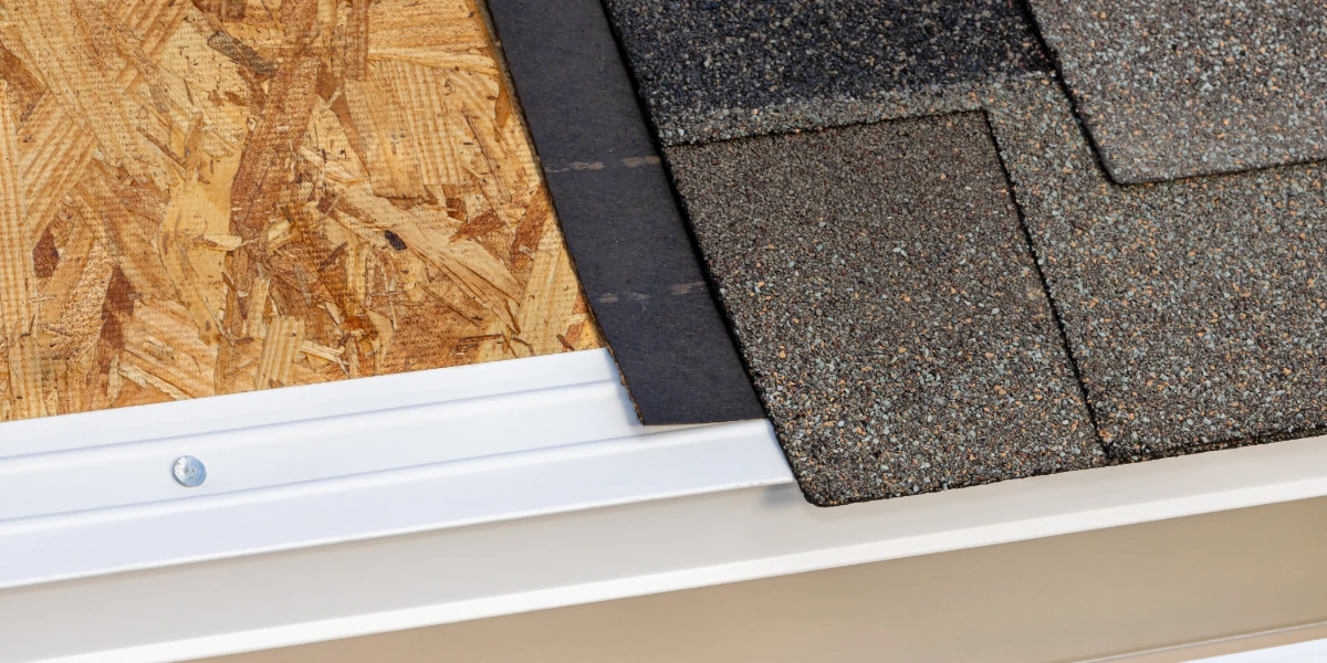 Close-up view of roof layers showing OSB decking, underlayment, asphalt shingles, and drip edge flashing on a residential roof.