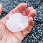 Hand holding a large hailstone over an asphalt shingle roof.